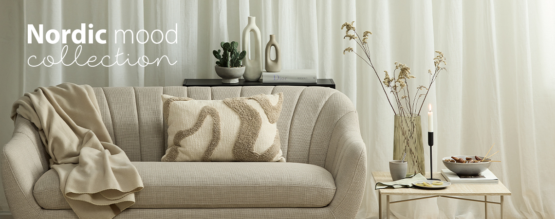Sofa mit Überwurf und getuftetem Kissen vor einer weißen Vase und neben einem Beistelltisch mit Vase, Kerzenhalter und Schale